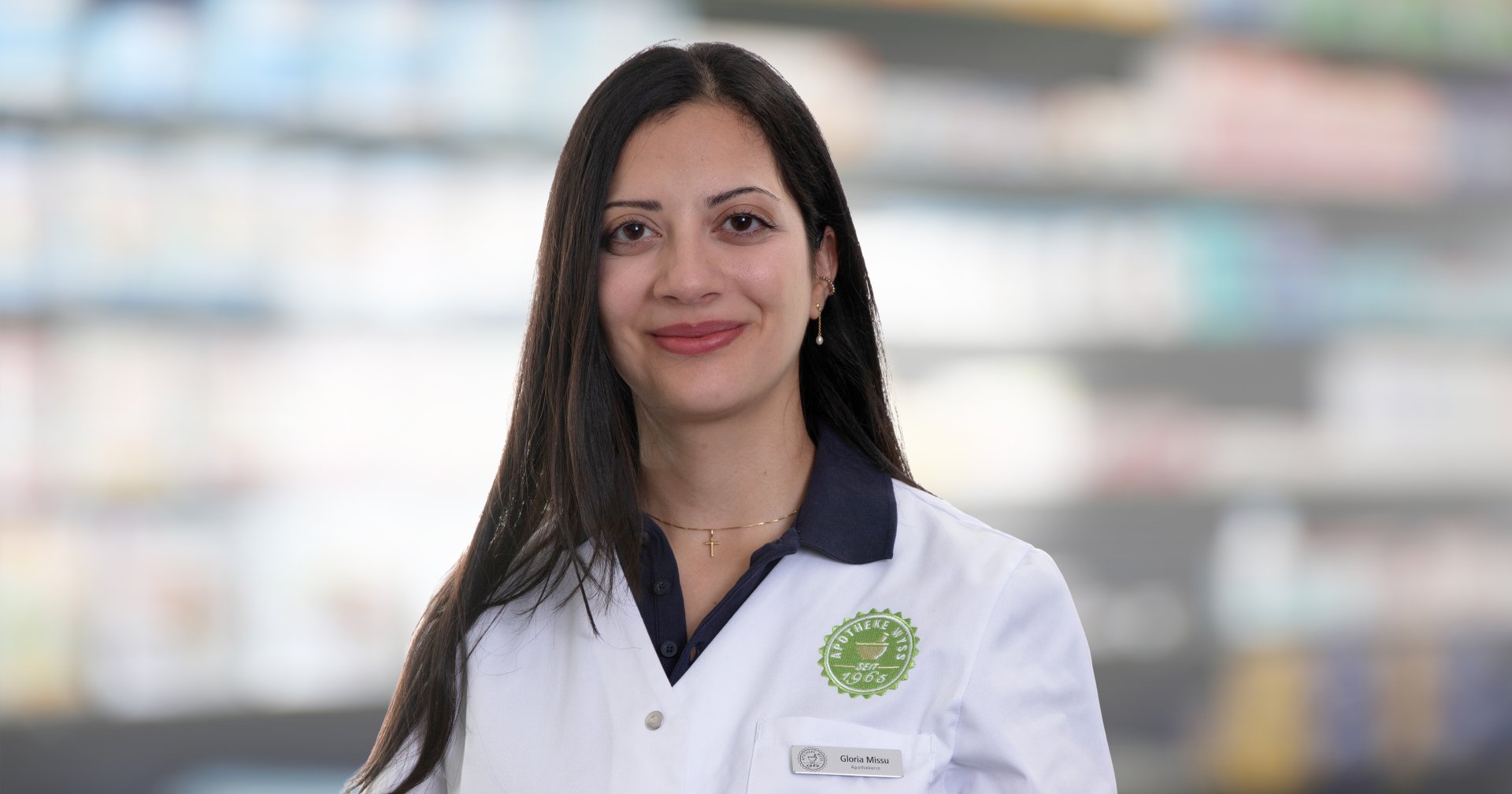 Joana Amaral Pedro, eine Frau mit langen dunklen Haaren, die einen wei&szlig;en Apothekerkittel tr&auml;gt, steht l&auml;chelnd vor verschwommenen Apothekenregalen.