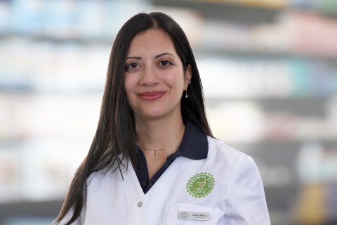 Joana Amaral Pedro, eine Frau mit langen dunklen Haaren, die einen wei&szlig;en Apothekerkittel tr&auml;gt, steht l&auml;chelnd vor verschwommenen Apothekenregalen.