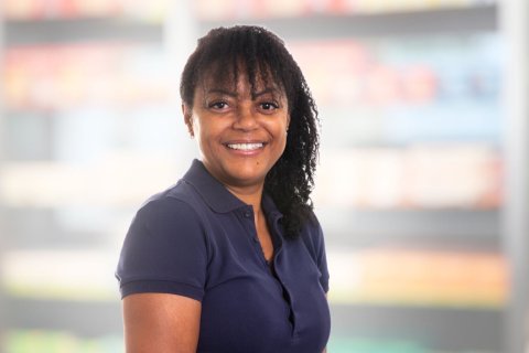 Joana Amaral Pedro, eine Frau mit lockigem Haar und einem marineblauen Poloshirt, steht l&auml;chelnd vor einem unscharfen Hintergrund, der Regale in einem Gesch&auml;ft oder einer Apotheke zu sein scheint.