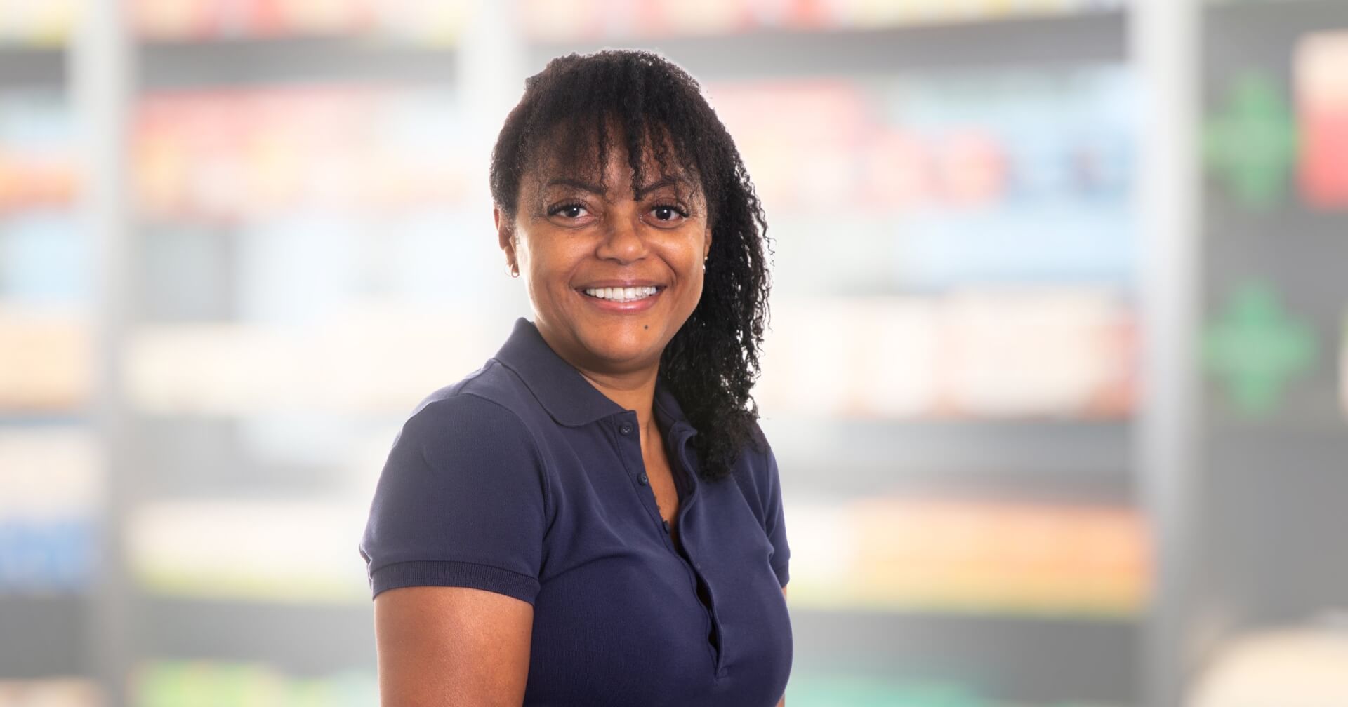 Joana Amaral Pedro, eine Frau mit lockigem Haar und einem marineblauen Poloshirt, steht l&auml;chelnd vor einem unscharfen Hintergrund, der Regale in einem Gesch&auml;ft oder einer Apotheke zu sein scheint.