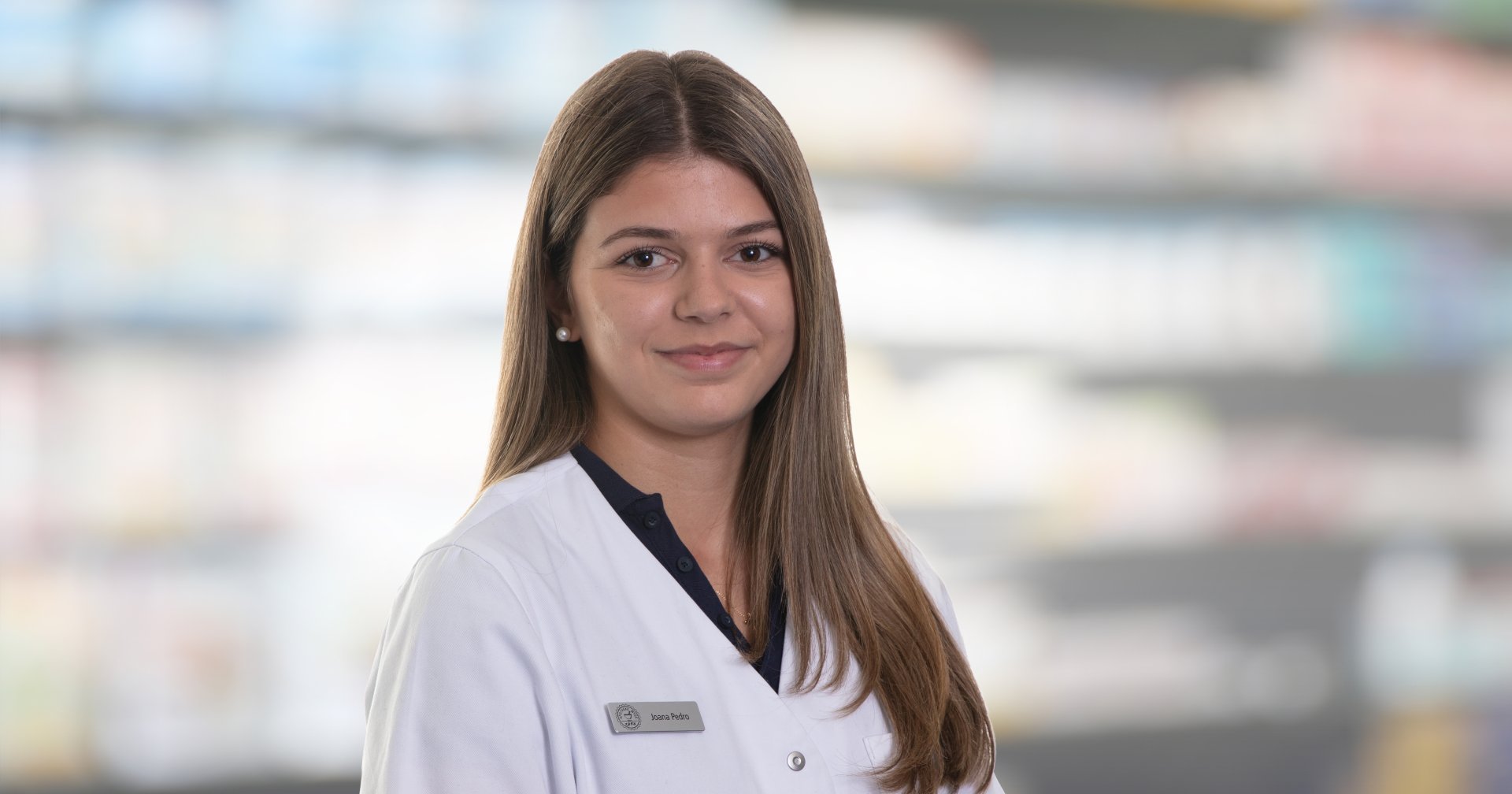 Joana Amaral Pedro, eine junge Frau mit langen braunen Haaren, tr&auml;gt einen wei&szlig;en Laborkittel und ein Namensschild, als sie l&auml;chelnd vor unscharfen Regalen in einer scheinbaren Apotheke oder medizinischen Einrichtung steht.