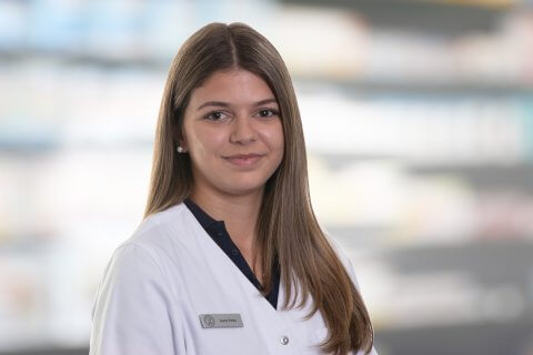 Joana Amaral Pedro, eine junge Frau mit langen braunen Haaren, tr&auml;gt einen wei&szlig;en Laborkittel und ein Namensschild, als sie l&auml;chelnd vor unscharfen Regalen in einer scheinbaren Apotheke oder medizinischen Einrichtung steht.