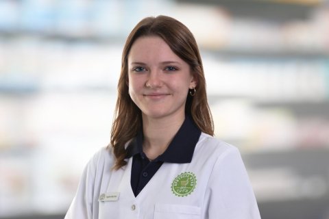 Joana Amaral Pedro, eine junge Frau mit wei&szlig;em Laborkittel und Namensschild, steht l&auml;chelnd vor den verschwommenen Apothekenregalen.