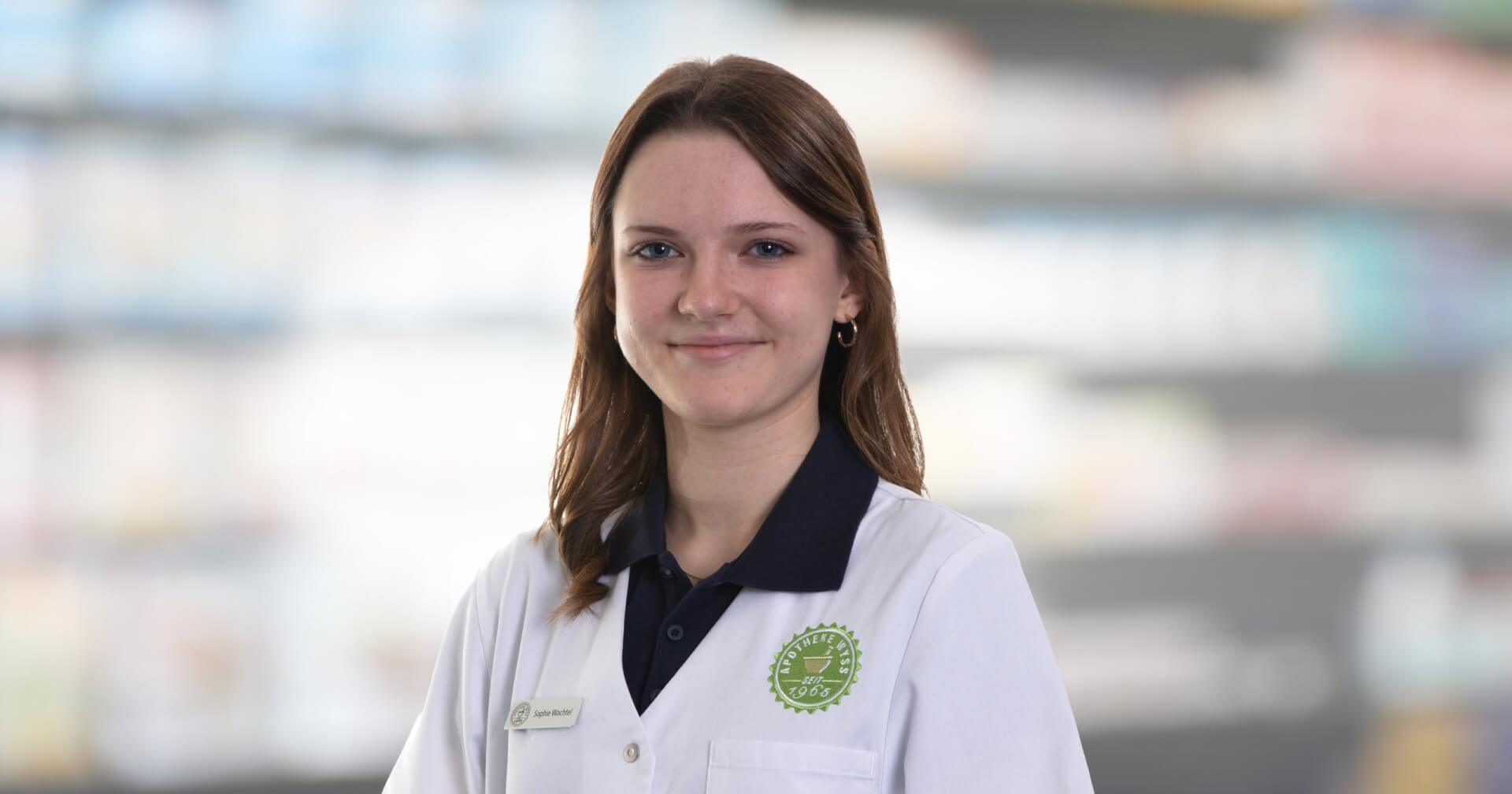 Joana Amaral Pedro, eine junge Frau mit wei&szlig;em Laborkittel und Namensschild, steht l&auml;chelnd vor den verschwommenen Apothekenregalen.