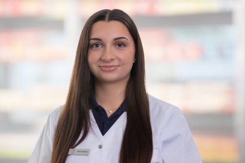 Joana Amaral Pedro, mit langen glatten braunen Haaren, wei&szlig;em Laborkittel und Namensschild, steht vor unscharfen Apothekenregalen. Sie schaut in die Kamera und l&auml;chelt leicht.