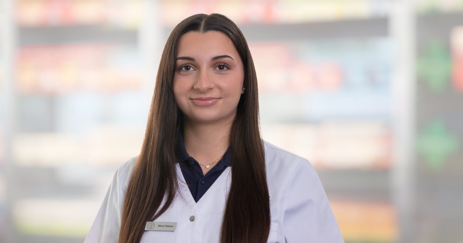 Joana Amaral Pedro, mit langen glatten braunen Haaren, wei&szlig;em Laborkittel und Namensschild, steht vor unscharfen Apothekenregalen. Sie schaut in die Kamera und l&auml;chelt leicht.