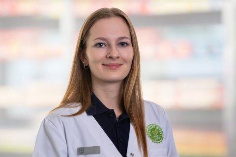 Eine junge Frau mit glattem, hellbraunem Haar, wei&szlig;em Kittel und dunklem Hemd, steht l&auml;chelnd vor den verschwommenen Apothekenregalen. Sie tr&auml;gt ein Namensschild und ein gr&uuml;nes Apothekenemblem auf ihrem Mantel.