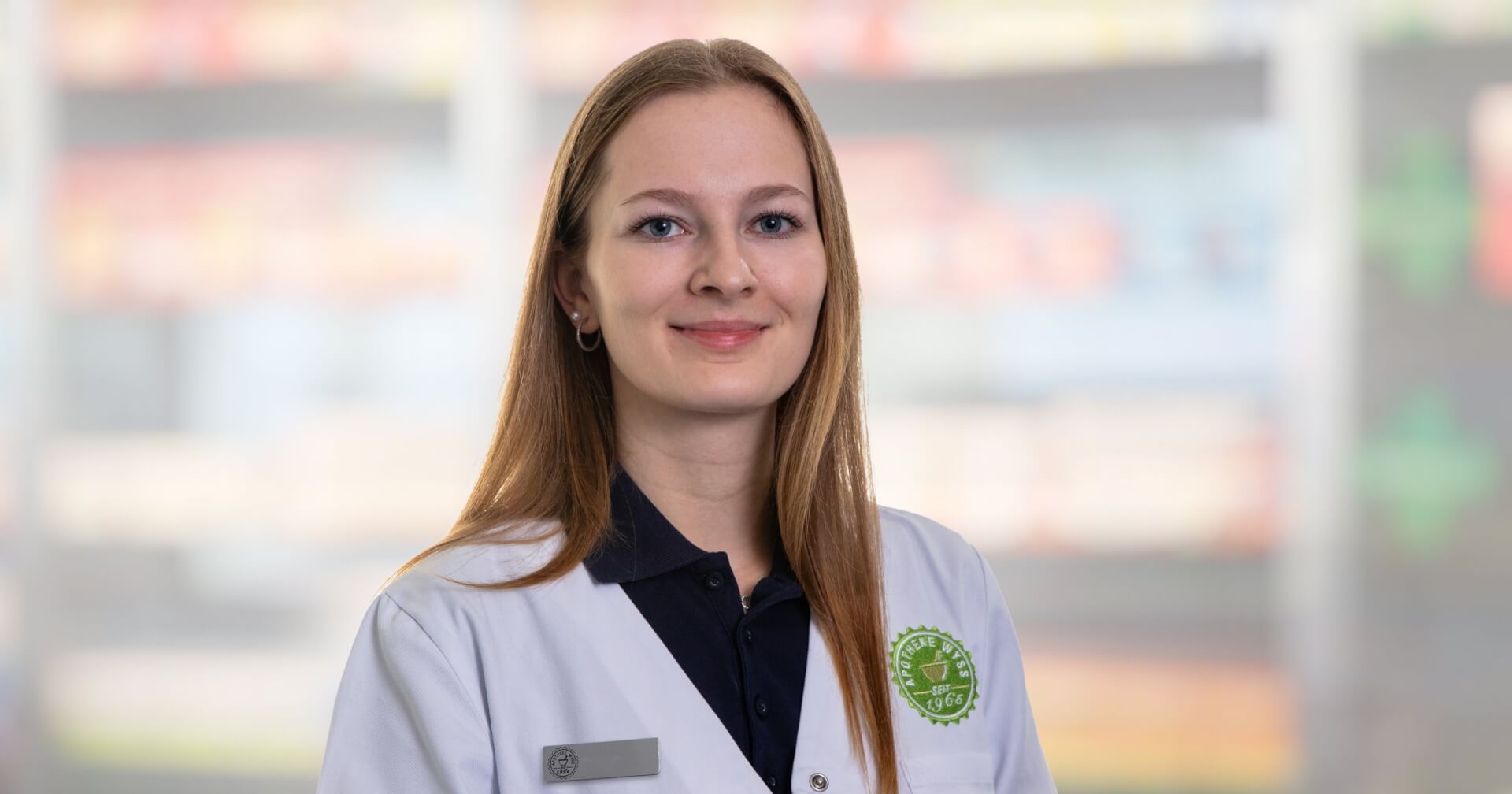 Eine junge Frau mit glattem, hellbraunem Haar, wei&szlig;em Kittel und dunklem Hemd, steht l&auml;chelnd vor den verschwommenen Apothekenregalen. Sie tr&auml;gt ein Namensschild und ein gr&uuml;nes Apothekenemblem auf ihrem Mantel.