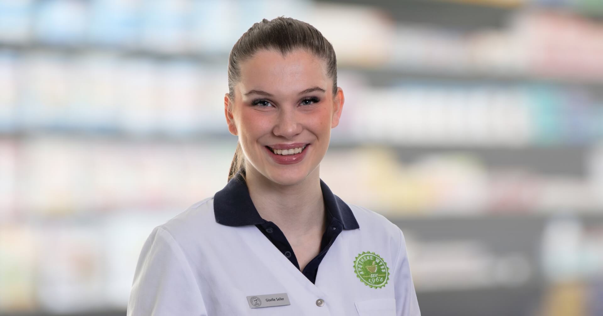 Eine l&auml;chelnde Frau, die durch ihr Namensschild als Joana Amaral Pedro identifiziert wird, tr&auml;gt einen wei&szlig;en Laborkittel, w&auml;hrend sie vor verschwommenen Apothekenregalen steht, was darauf hindeutet, dass sie Apothekerin oder medizinisches Fachpersonal ist.