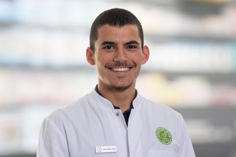 Ein junger Mann, der einen wei&szlig;en Laborkittel mit einem gr&uuml;nen Emblem und einem Namensschild mit der Aufschrift Joana Amaral Pedro tr&auml;gt, l&auml;chelt in die Kamera. Der unscharfe Hintergrund deutet auf einen Innenraum hin, m&ouml;glicherweise eine Apotheke oder ein Labor.