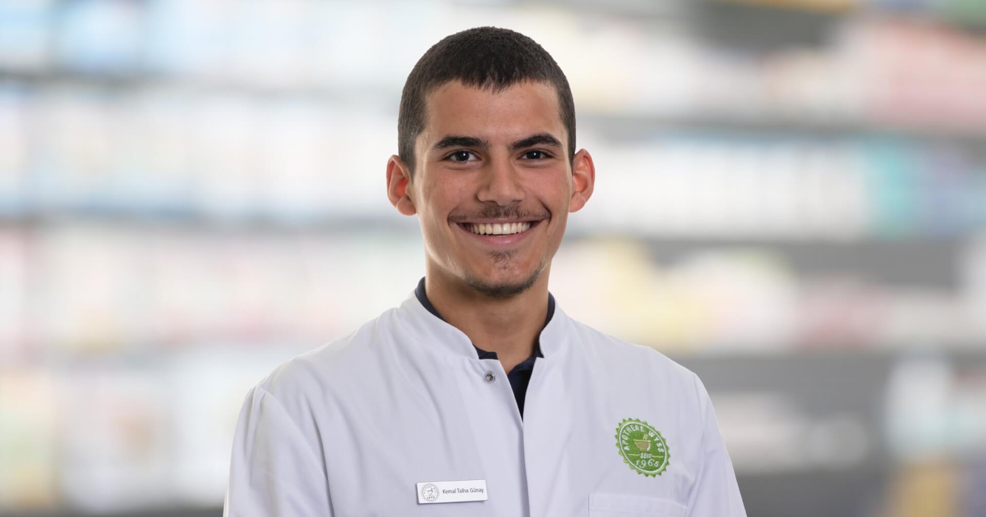 Ein junger Mann, der einen wei&szlig;en Laborkittel mit einem gr&uuml;nen Emblem und einem Namensschild mit der Aufschrift Joana Amaral Pedro tr&auml;gt, l&auml;chelt in die Kamera. Der unscharfe Hintergrund deutet auf einen Innenraum hin, m&ouml;glicherweise eine Apotheke oder ein Labor.