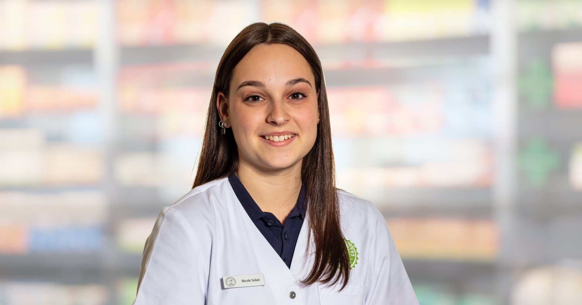 Nicole Solari, eine junge Frau in einem wei&szlig;en Laborkittel mit Namensschild, steht l&auml;chelnd vor verschwommenen Regalen, m&ouml;glicherweise in einer Apotheke oder einem Gesch&auml;ft.