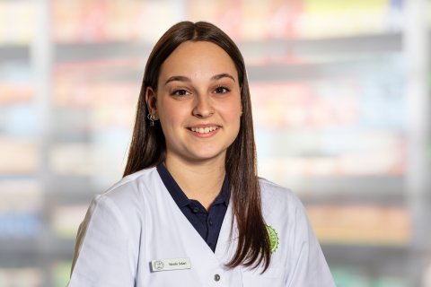 Nicole Solari, eine junge Frau in einem wei&szlig;en Laborkittel mit Namensschild, steht l&auml;chelnd vor verschwommenen Regalen, m&ouml;glicherweise in einer Apotheke oder einem Gesch&auml;ft.