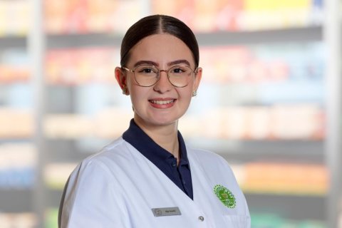 Eine l&auml;chelnde Frau mit Brille und wei&szlig;em Laborkittel steht vor verschwommenen Regalen, m&ouml;glicherweise in einer Apotheke oder einem Gesch&auml;ft. Sie hat ihr dunkles Haar zur&uuml;ckgebunden und tr&auml;gt ein Namensschild an ihrem Kittel.