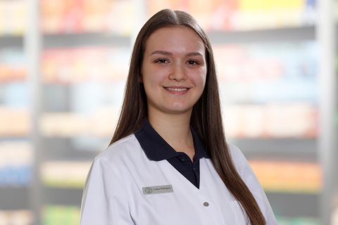 Eine junge Frau mit langen braunen Haaren, die einen wei&szlig;en Laborkittel mit einem Namensschild tr&auml;gt, auf dem "Luana Rodrigues" steht, l&auml;chelt in die Kamera. Der unscharfe Hintergrund, der mit Regalen voller Produkte gef&uuml;llt ist, deutet auf eine Apotheke oder ein Gesch&auml;ft hin.