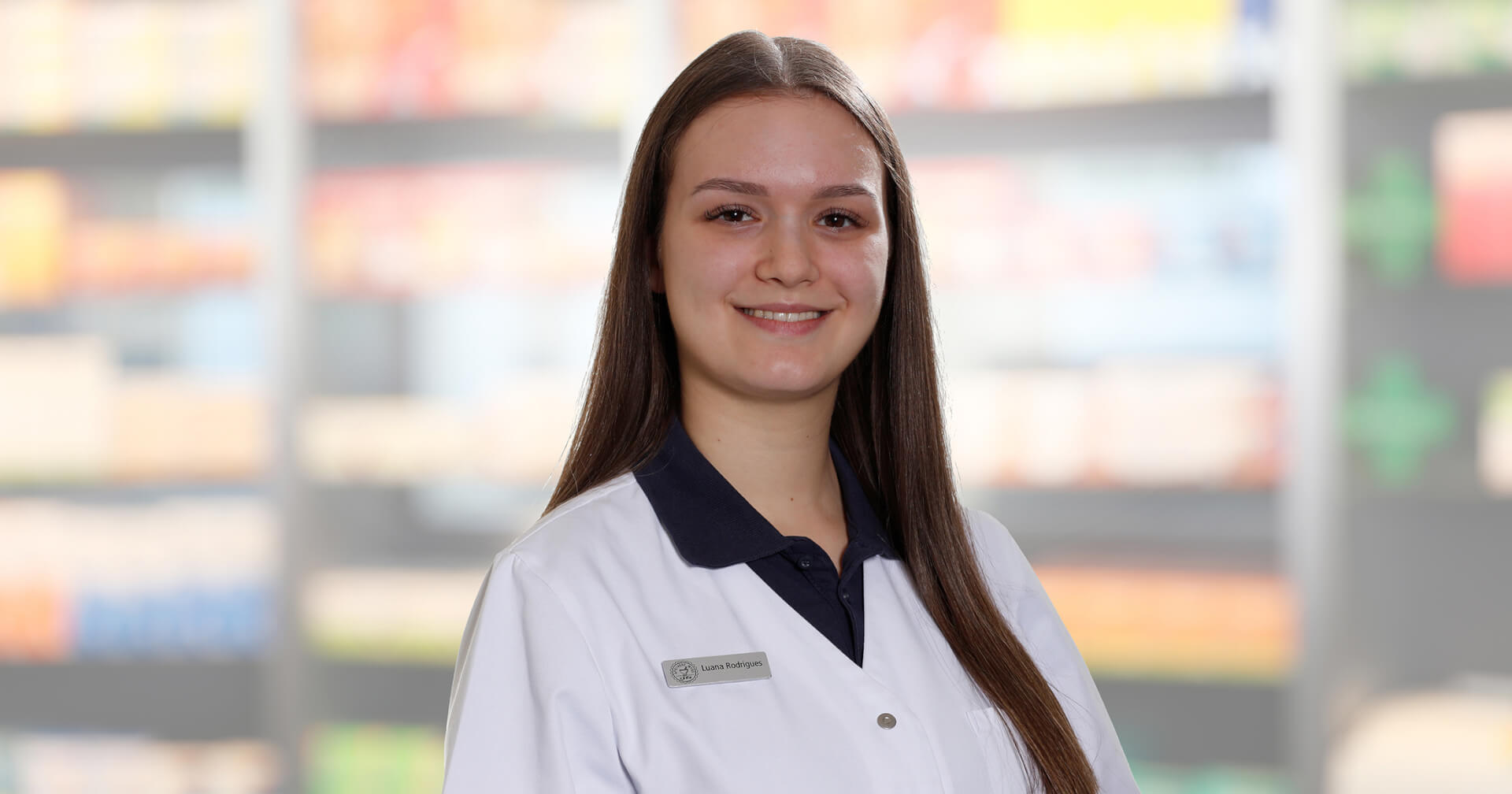 Eine junge Frau mit langen braunen Haaren, die einen wei&szlig;en Laborkittel mit einem Namensschild tr&auml;gt, auf dem "Luana Rodrigues" steht, l&auml;chelt in die Kamera. Der unscharfe Hintergrund, der mit Regalen voller Produkte gef&uuml;llt ist, deutet auf eine Apotheke oder ein Gesch&auml;ft hin.