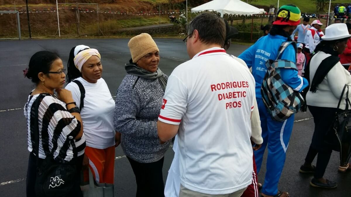 Eine Gruppe von Menschen unterhält sich im Freien; ein Mann in einem weißen Hilfswerk Rodrigues Diabetic Pros Shirt spricht mit drei Frauen, von denen eine einen beigen Hut trägt. Andere in bunter Kleidung stehen im Hintergrund auf einer gepflasterten Fläche.