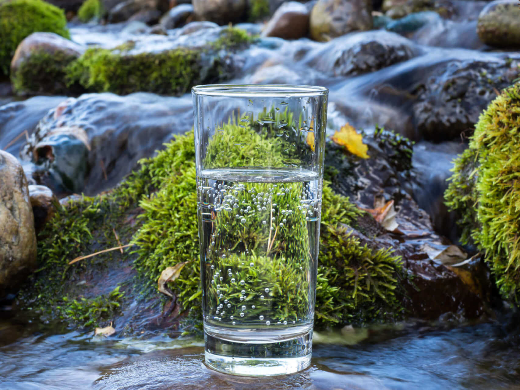Ein klares Wasserglas steht auf moosbewachsenen Felsen in einem flie&szlig;enden Bach, in dessen Oberfl&auml;che sich das gr&uuml;ne Moos und die umgebende Natur spiegeln und eine nat&uuml;rliche, heitere Szene schaffen.
