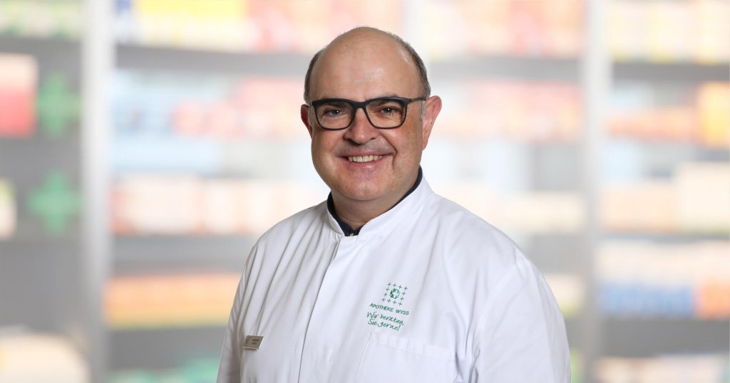 Ein l&auml;chelnder Mann mit Brille und wei&szlig;em Laborkittel steht vor verschwommenen Regalen, wahrscheinlich in einer Apotheke oder in einem medizinischen Bereich.