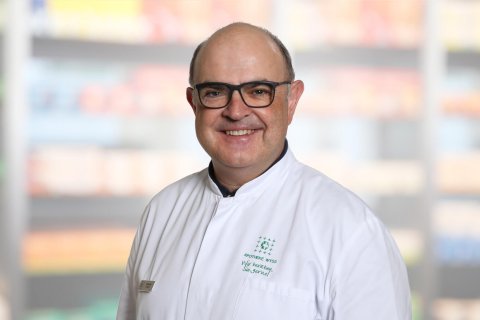 Ein l&auml;chelnder Mann mit Brille und wei&szlig;em Laborkittel steht vor verschwommenen Regalen, wahrscheinlich in einer Apotheke oder in einem medizinischen Bereich.