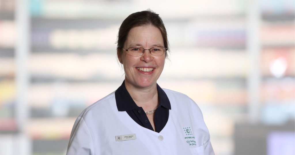 Barbara G&ouml;ring, eine l&auml;chelnde Frau mit Brille und wei&szlig;em Laborkittel, steht vor einem unscharfen Hintergrund. Sie hat ihr braunes Haar zur&uuml;ckgebunden und tr&auml;gt unter dem Laborkittel ein dunkles Hemd mit Kragen.