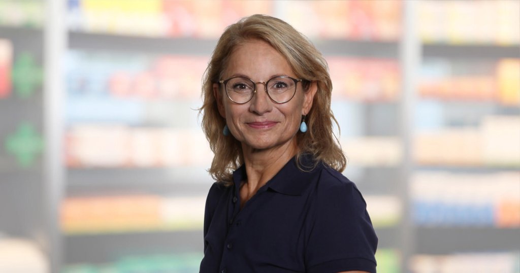 Helen Hagmann, eine Frau mittleren Alters mit blondem Haar, Brille und blauen Ohrringen, l&auml;chelt und tr&auml;gt ein marineblaues Poloshirt. Sie steht vor verschwommenen Regalen, m&ouml;glicherweise in einem Gesch&auml;ft oder einer Apotheke.