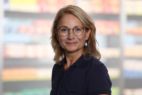 Helen Hagmann, eine Frau mittleren Alters mit blondem Haar, Brille und blauen Ohrringen, l&auml;chelt und tr&auml;gt ein marineblaues Poloshirt. Sie steht vor verschwommenen Regalen, m&ouml;glicherweise in einem Gesch&auml;ft oder einer Apotheke.