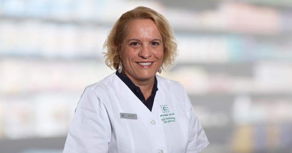 Milica Da&scaron;ić, eine Frau mit blondem Haar, l&auml;chelt, w&auml;hrend sie einen wei&szlig;en Laborkittel und ein Namensschild tr&auml;gt. Sie steht vor unscharfen Regalen, wahrscheinlich in einer Apotheke oder einem medizinischen Bereich.