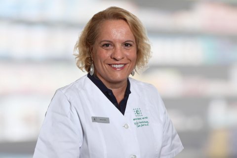 Milica Da&scaron;ić, eine Frau mit blondem Haar, l&auml;chelt, w&auml;hrend sie einen wei&szlig;en Laborkittel und ein Namensschild tr&auml;gt. Sie steht vor unscharfen Regalen, wahrscheinlich in einer Apotheke oder einem medizinischen Bereich.