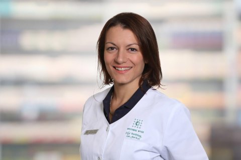 Eine Frau in einem wei&szlig;en Laborkittel l&auml;chelt, w&auml;hrend sie in einem Innenraum vor einem unscharfen Hintergrund steht, der eine professionelle oder medizinische Umgebung andeutet. Antonella Celestre strahlt in ihrer Rolle Zuversicht und Herzlichkeit aus.