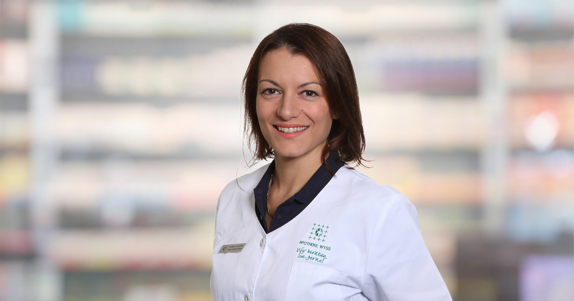 Eine Frau in einem wei&szlig;en Laborkittel l&auml;chelt, w&auml;hrend sie in einem Innenraum vor einem unscharfen Hintergrund steht, der eine professionelle oder medizinische Umgebung andeutet. Antonella Celestre strahlt in ihrer Rolle Zuversicht und Herzlichkeit aus.