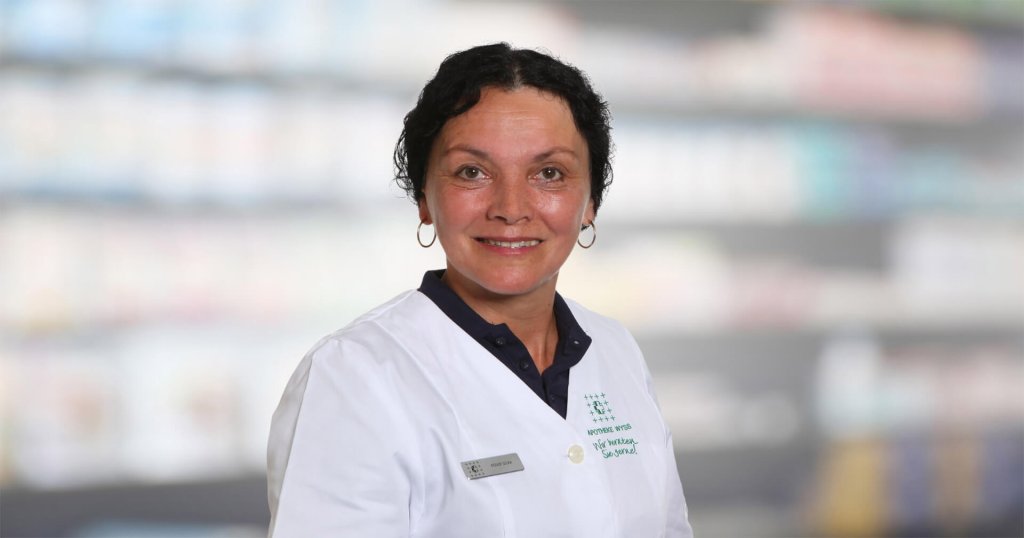 Silvia R&ouml;der, eine Frau mit kurzen dunklen Haaren, die einen wei&szlig;en Laborkittel und ein Namensschild tr&auml;gt, l&auml;chelt in die Kamera. Der unscharfe Hintergrund deutet auf Regale oder eine Apothekeneinrichtung hin.