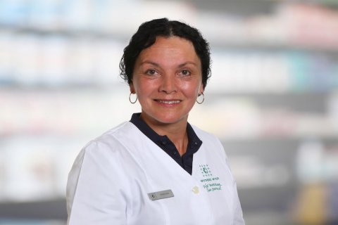 Silvia R&ouml;der, eine Frau mit kurzen dunklen Haaren, die einen wei&szlig;en Laborkittel und ein Namensschild tr&auml;gt, l&auml;chelt in die Kamera. Der unscharfe Hintergrund deutet auf Regale oder eine Apothekeneinrichtung hin.