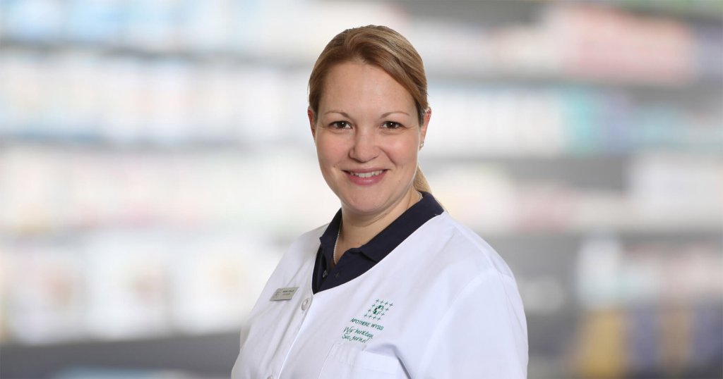 Mariana Schaller, eine l&auml;chelnde Frau in einem wei&szlig;en Laborkittel, steht vor verschwommenen Regalen, die an eine Apotheke oder einen medizinischen Bereich erinnern.