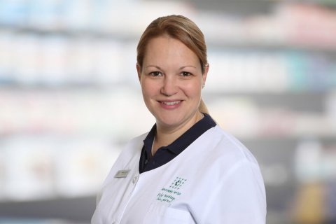 Mariana Schaller, eine l&auml;chelnde Frau in einem wei&szlig;en Laborkittel, steht vor verschwommenen Regalen, die an eine Apotheke oder einen medizinischen Bereich erinnern.