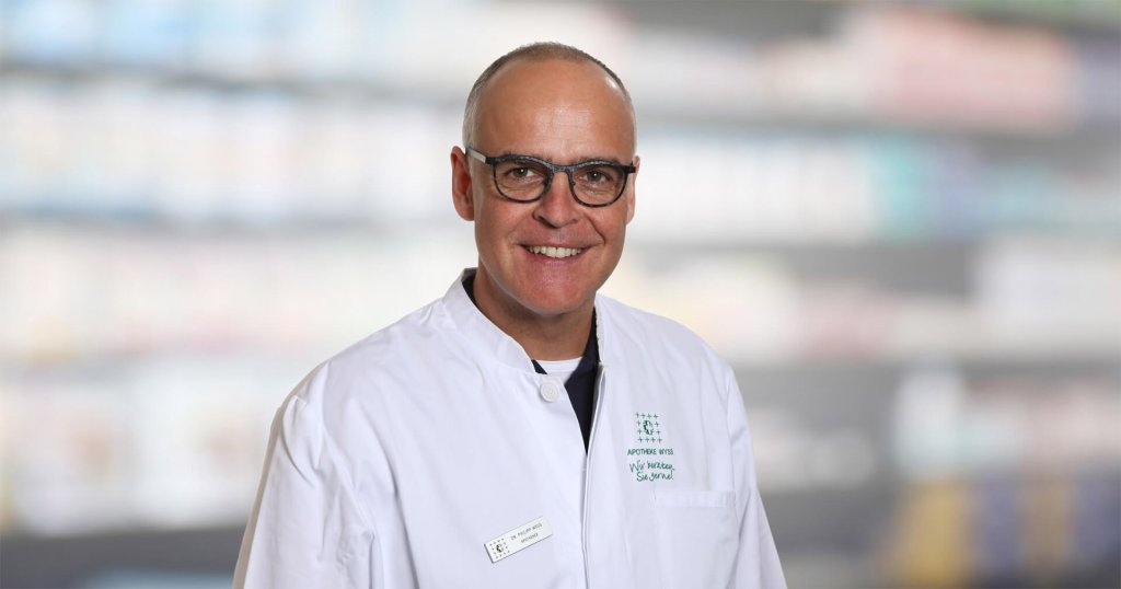 Dr. Philipp Wyss, ein l&auml;chelnder Mann mit Brille und wei&szlig;em Laborkittel, steht vor unscharfen Regalen, m&ouml;glicherweise in einer Apotheke oder einem medizinischen Bereich.