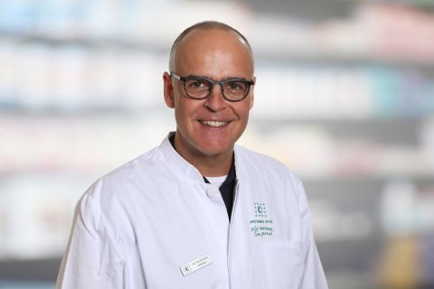 Dr. Philipp Wyss, ein l&auml;chelnder Mann mit Brille und wei&szlig;em Laborkittel, steht vor unscharfen Regalen, m&ouml;glicherweise in einer Apotheke oder einem medizinischen Bereich.