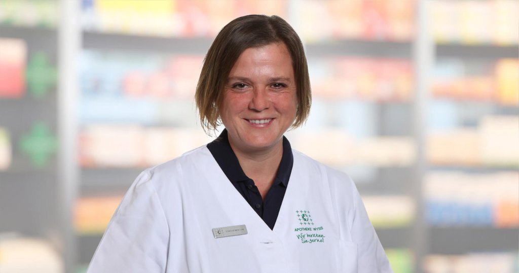 Natascha Reinhard, eine Frau mit kurzen braunen Haaren, die einen wei&szlig;en Laborkittel und ein Namensschild tr&auml;gt, l&auml;chelt, w&auml;hrend sie vor unscharfen Regalen steht, m&ouml;glicherweise in einer Apotheke oder einer medizinischen Einrichtung.