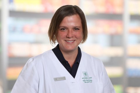 Natascha Reinhard, eine Frau mit kurzen braunen Haaren, die einen wei&szlig;en Laborkittel und ein Namensschild tr&auml;gt, l&auml;chelt, w&auml;hrend sie vor unscharfen Regalen steht, m&ouml;glicherweise in einer Apotheke oder einer medizinischen Einrichtung.