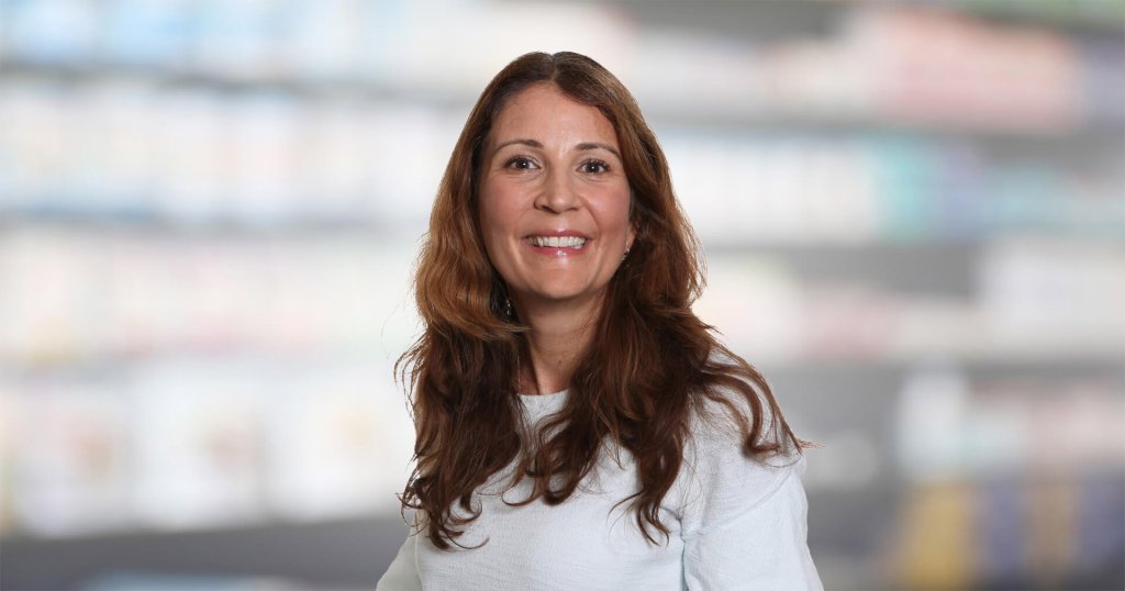 Magdalena Doukas, eine Frau mit langen braunen Haaren und einem wei&szlig;en Oberteil, l&auml;chelt in die Kamera, w&auml;hrend sie vor einem unscharfen Innenhintergrund mit Regalen steht.