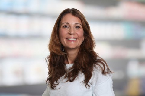 Magdalena Doukas, eine Frau mit langen braunen Haaren und einem wei&szlig;en Oberteil, l&auml;chelt in die Kamera, w&auml;hrend sie vor einem unscharfen Innenhintergrund mit Regalen steht.