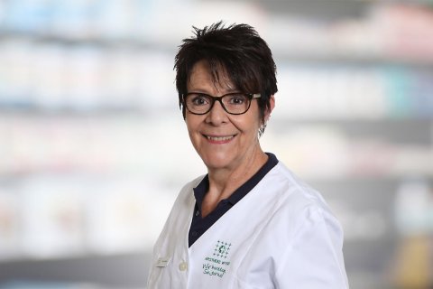 Barbara Zmoos, eine Frau mit kurzen dunklen Haaren und Brille, l&auml;chelt, w&auml;hrend sie einen wei&szlig;en Laborkittel tr&auml;gt und vor einem unscharfen Hintergrund steht, der Regale in einer Apotheke oder einem Gesch&auml;ft darzustellen scheint.