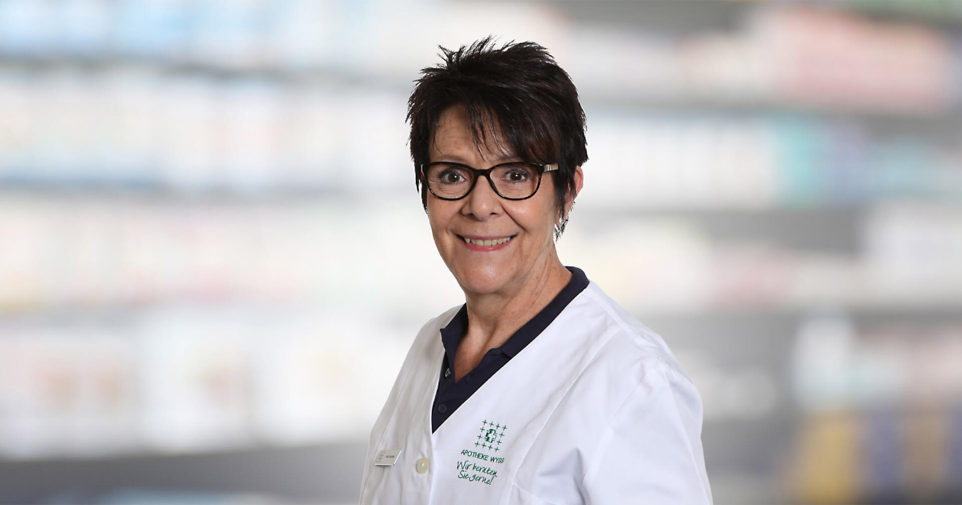 Barbara Zmoos, eine Frau mit kurzen dunklen Haaren und Brille, l&auml;chelt, w&auml;hrend sie einen wei&szlig;en Laborkittel tr&auml;gt und vor einem unscharfen Hintergrund steht, der Regale in einer Apotheke oder einem Gesch&auml;ft darzustellen scheint.