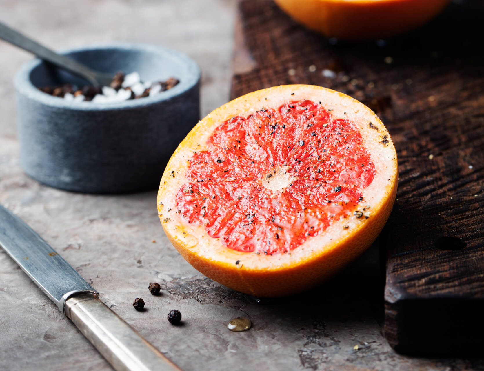 Eine halbierte, bittere und doch gesunde Grapefruit, bestreut mit Pfeffer, liegt auf einer rustikalen Fl&auml;che neben einem Holzbrett, einem Messer, Pfefferk&ouml;rnern und einer kleinen Schale mit Salz und einem L&ouml;ffel.