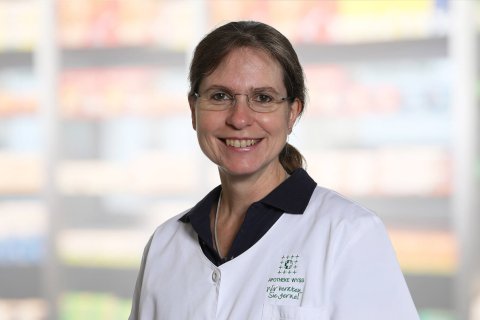 Eine Frau mit einer Brille und einem wei&szlig;en Laborkittel l&auml;chelt in die Kamera. Der Hintergrund ist unscharf und zeigt Regale mit bunten Produkten.