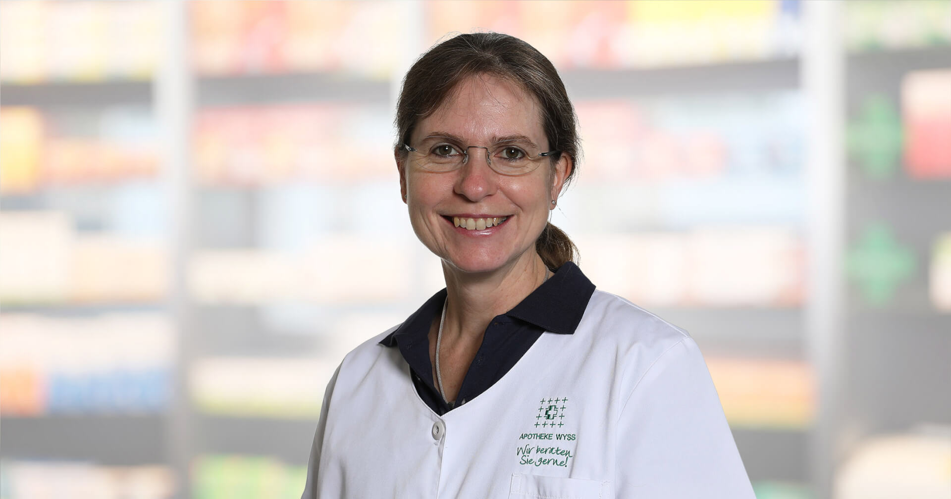 Eine Frau mit einer Brille und einem wei&szlig;en Laborkittel l&auml;chelt in die Kamera. Der Hintergrund ist unscharf und zeigt Regale mit bunten Produkten.