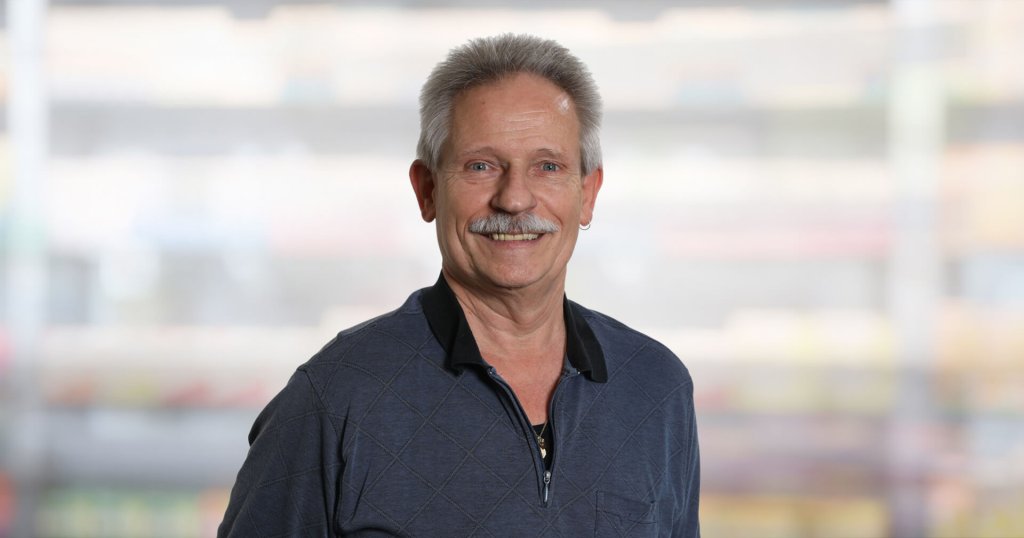 Hanspeter K&auml;gi, ein Mann mittleren Alters mit kurzen grauen Haaren und einem Schnurrbart, l&auml;chelt in die Kamera. Er tr&auml;gt einen dunklen, gemusterten Pullover mit Rei&szlig;verschluss &uuml;ber einem schwarzen Hemd und steht vor einem unscharfen, hellen Hintergrund.