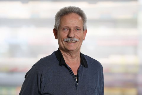 Hanspeter K&auml;gi, ein Mann mittleren Alters mit kurzen grauen Haaren und einem Schnurrbart, l&auml;chelt in die Kamera. Er tr&auml;gt einen dunklen, gemusterten Pullover mit Rei&szlig;verschluss &uuml;ber einem schwarzen Hemd und steht vor einem unscharfen, hellen Hintergrund.