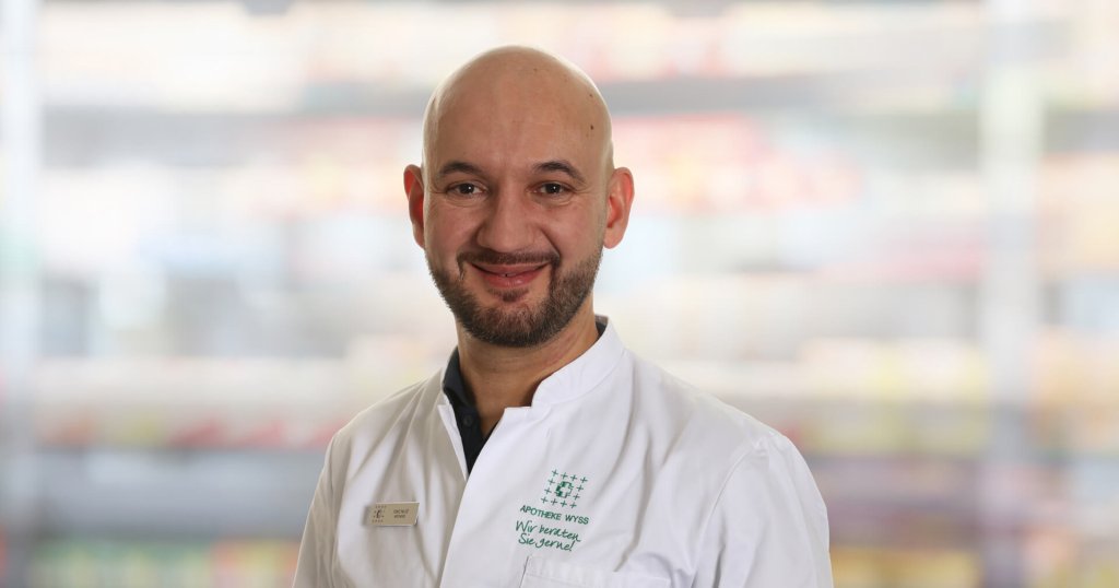 Ein l&auml;chelnder Mann mit Glatze und gestutztem Bart tr&auml;gt einen wei&szlig;en Arztkittel und steht vor einem unscharfen Hintergrund mit Regalen.