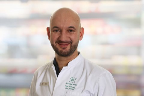 Ein l&auml;chelnder Mann mit Glatze und gestutztem Bart tr&auml;gt einen wei&szlig;en Arztkittel und steht vor einem unscharfen Hintergrund mit Regalen.